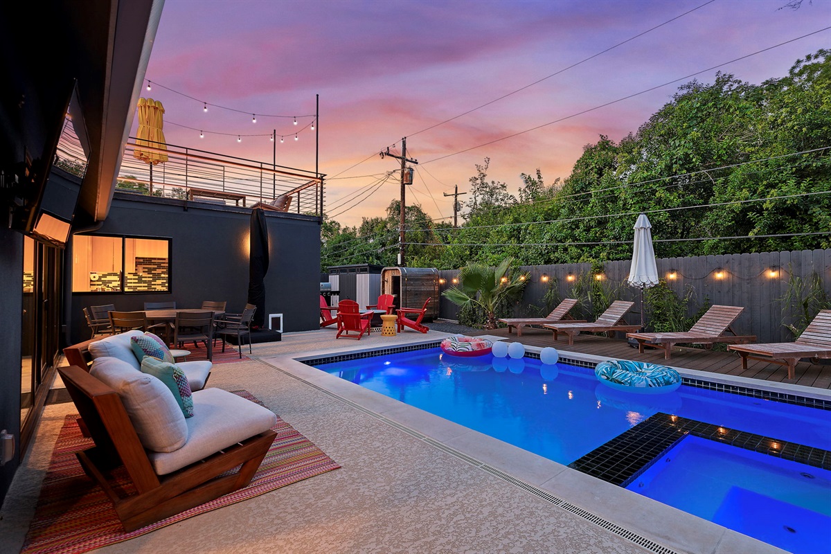 A vibrant backyard oasis at dusk, featuring a sparkling blue swimming pool, multiple lounging and dining areas with string lights, and a second-story deck, all enclosed by a modern wooden fence.