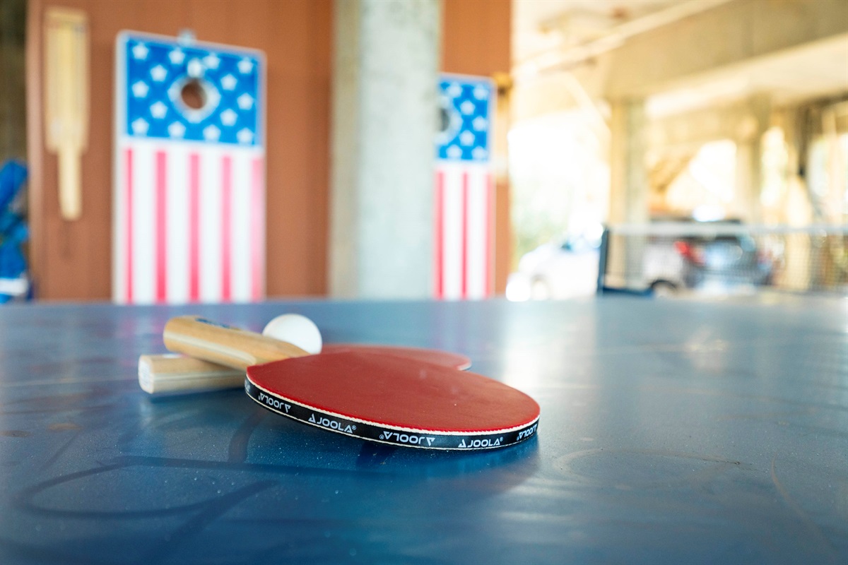 Enjoy a casual ping pong match with JOOLA gear on a vibrant blue table and festive cornhole boards nearby.
