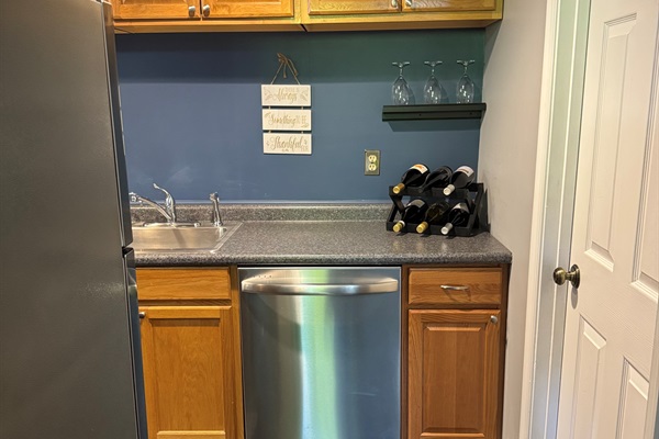 Kitchen area with dishwasher and a variety of bottled wine with drinking glasses.