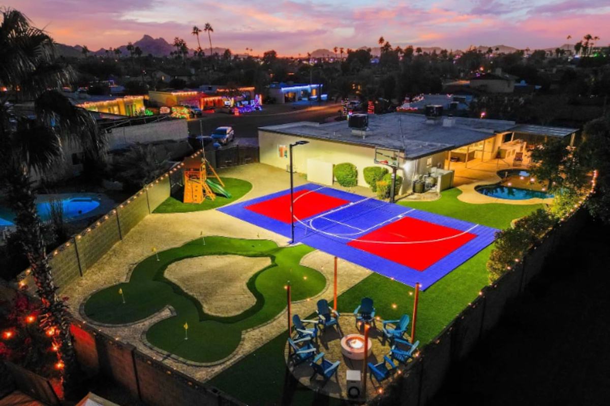 As the lights come on, the whole backyard turns into one big invitation — court, green, fire pit lounge, and string lights overhead.