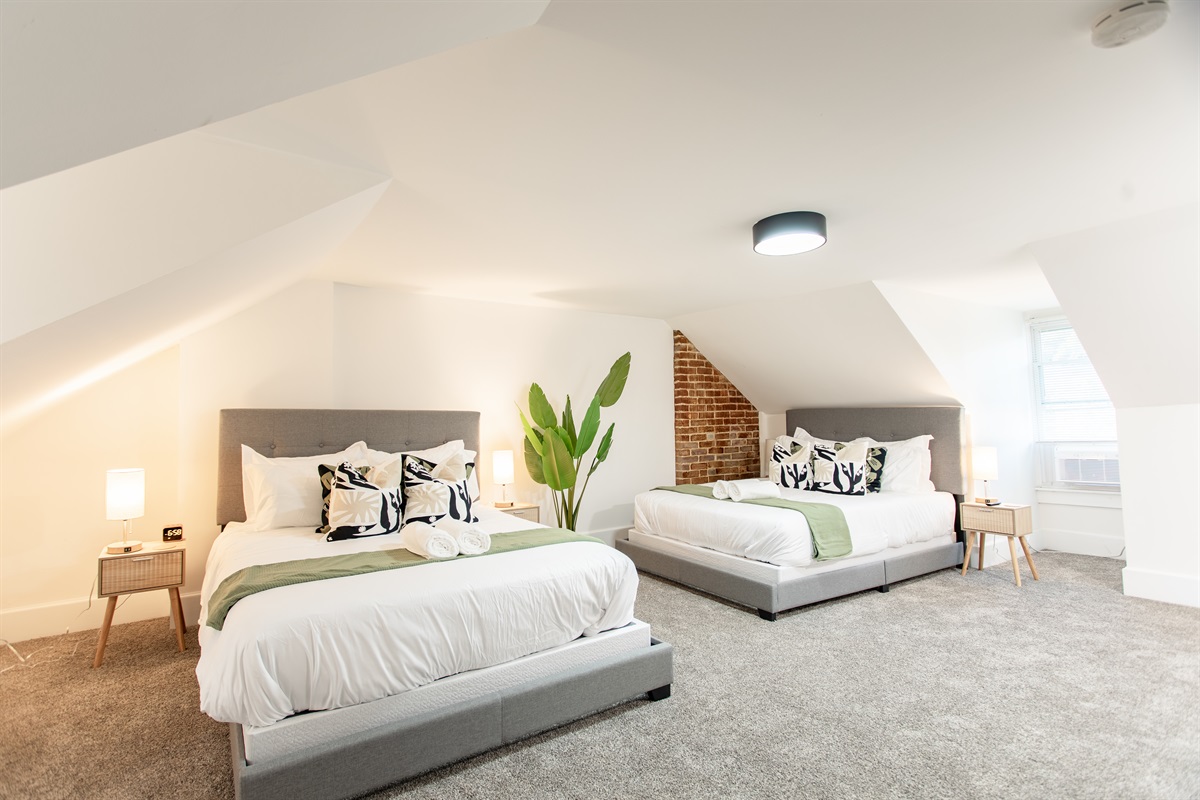 Spacious top-floor bedroom with two queen beds, modern décor, and exposed brick accents, complemented by a dedicated workspace for added convenience.