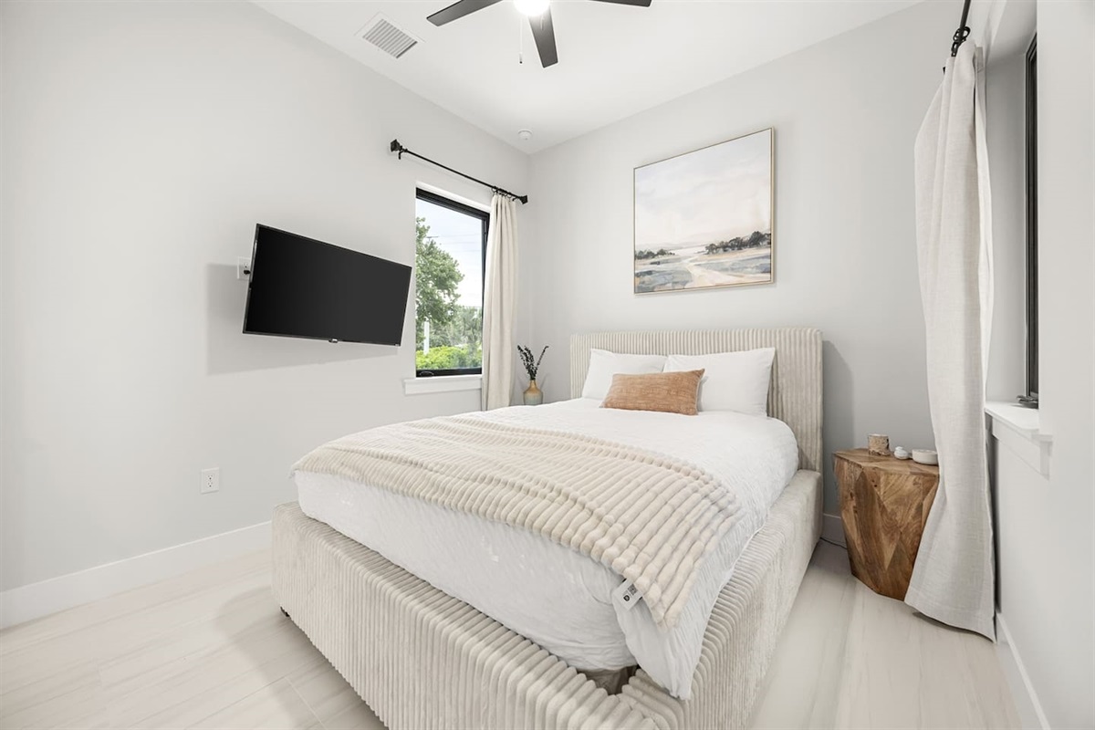 First floor queen bedroom with a light and airy design, featuring soft decor, a ceiling fan for comfort, and a TV for relaxation.