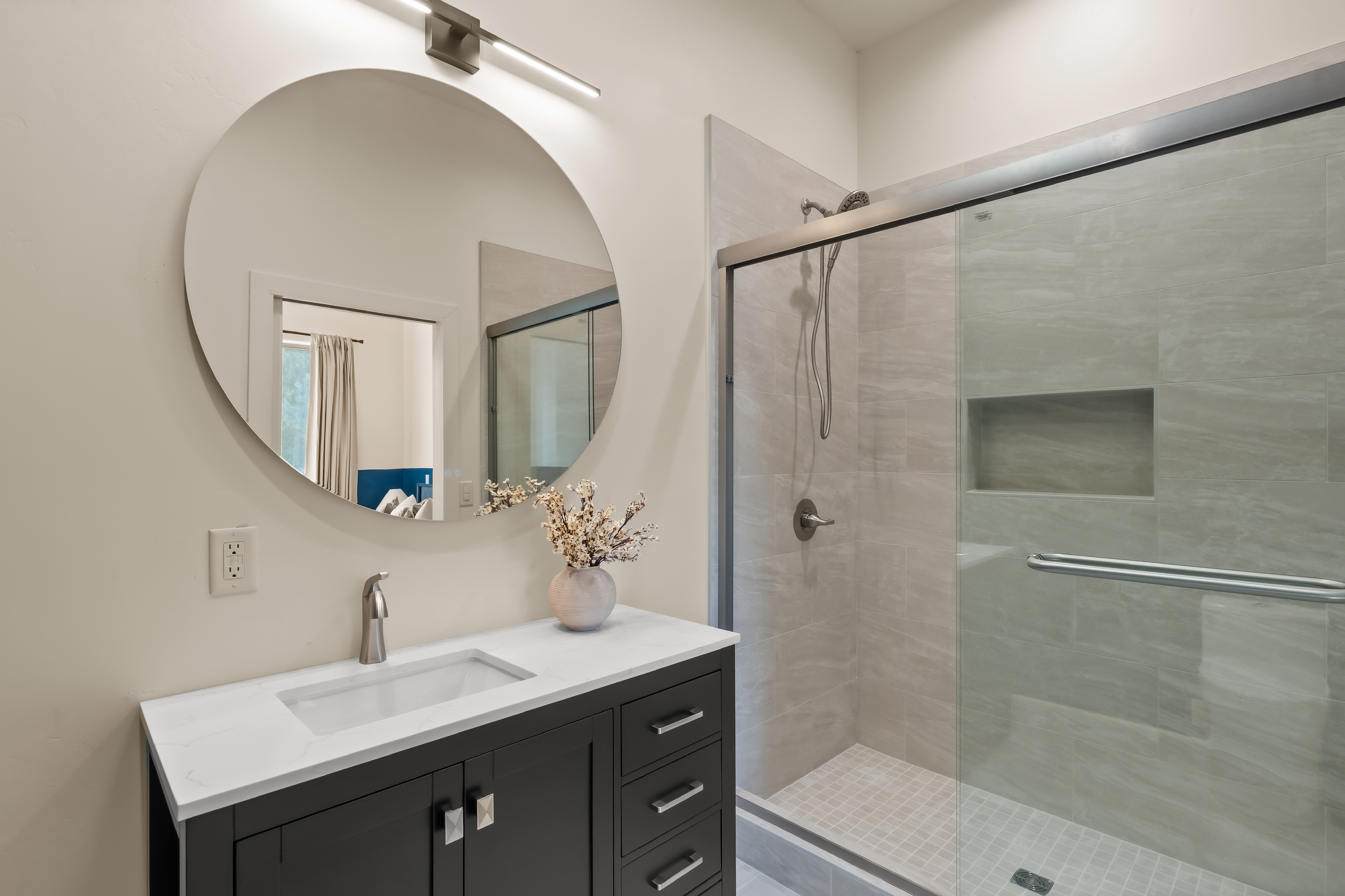 Modern ensuite bath featuring a sleek black vanity, bright round mirror, and a spacious glass-enclosed walk-in tile shower with built-in wall niche.