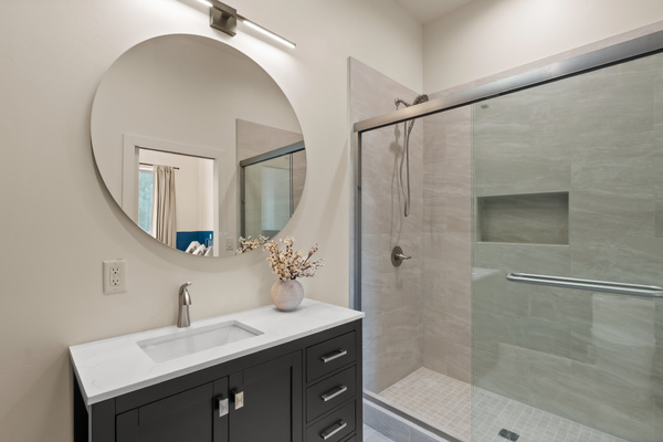 Modern ensuite bath featuring a sleek black vanity, bright round mirror, and a spacious glass-enclosed walk-in tile shower with built-in wall niche.