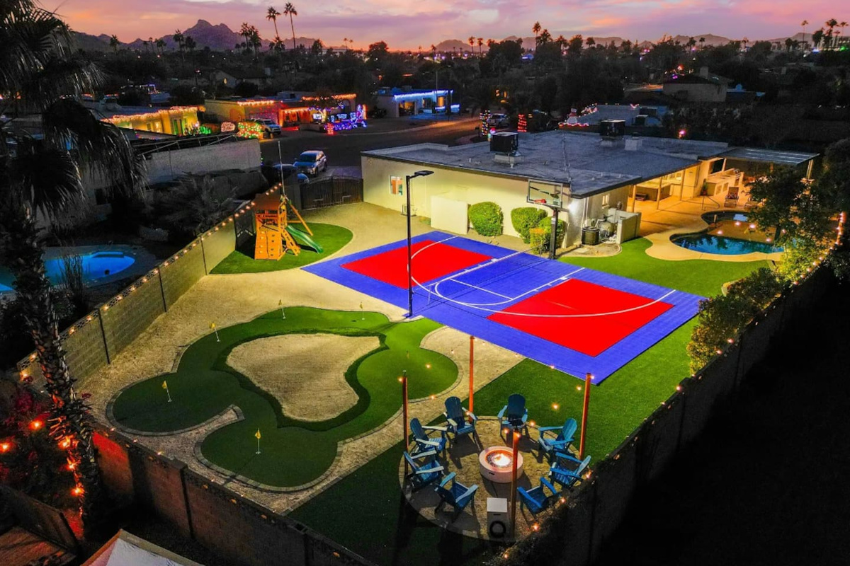 As the lights come on, the whole backyard turns into one big invitation — court, green, fire pit lounge, and string lights overhead.