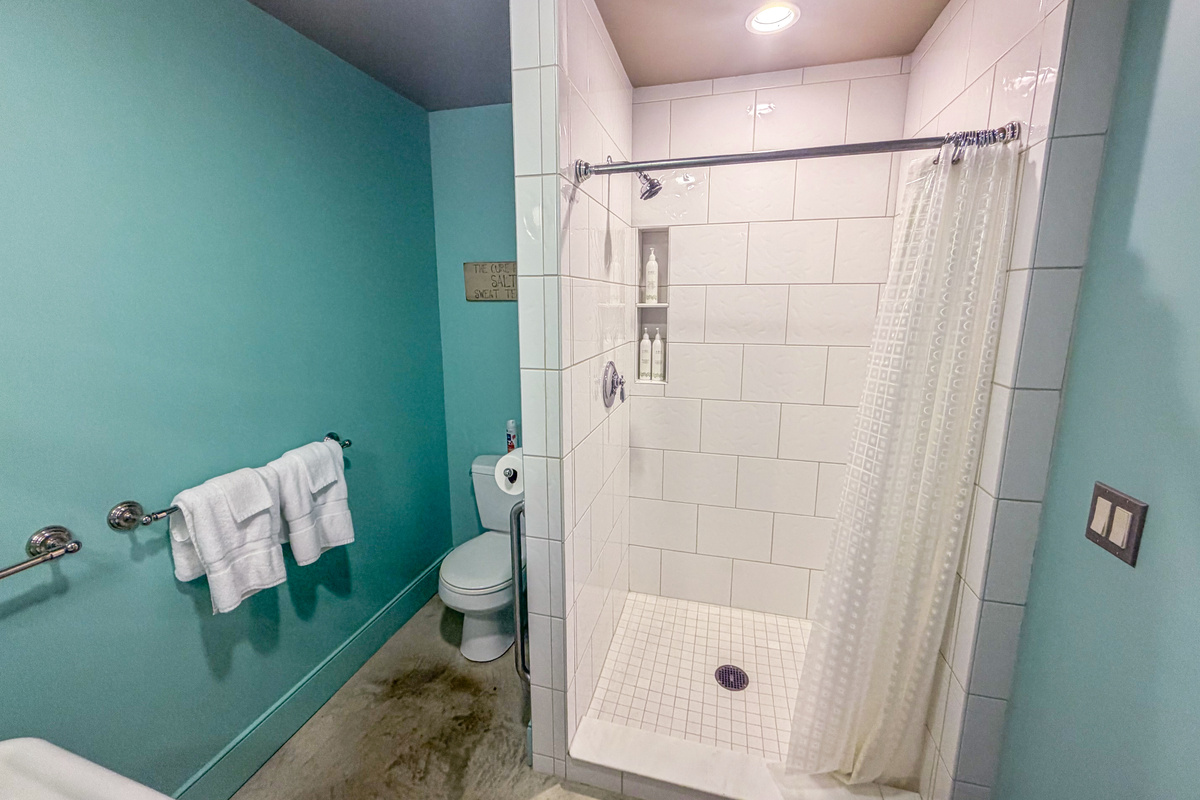 Inside the Base Camp full bath: a spacious step-in shower with classic white tile, built-in shampoo niches, polished concrete flooring, and fresh linens. Complete private bath convenience on "The Crag's" ground floor.