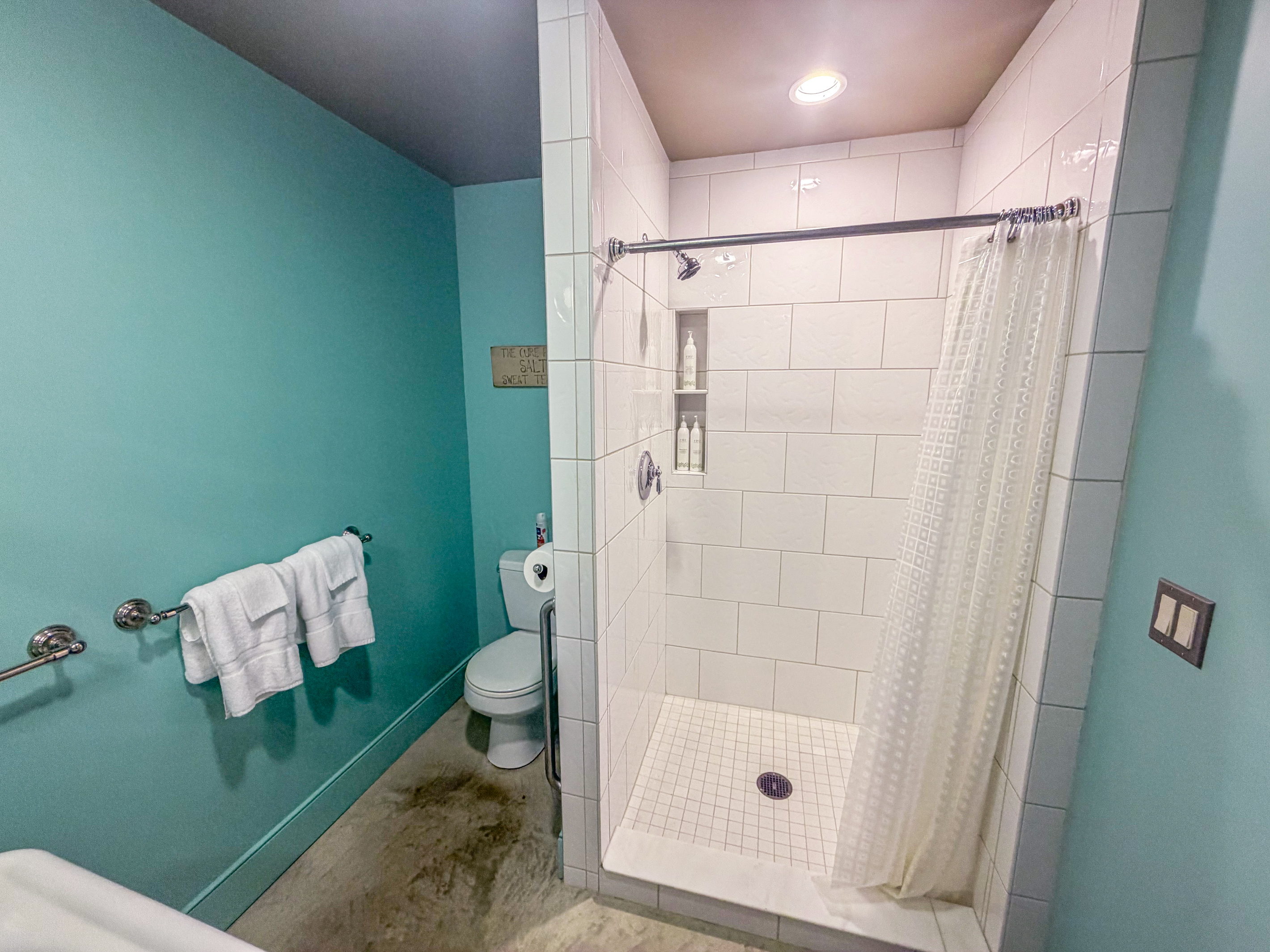 Inside the Base Camp full bath: a spacious step-in shower with classic white tile, built-in shampoo niches, polished concrete flooring, and fresh linens. Complete private bath convenience on "The Crag's" ground floor.
