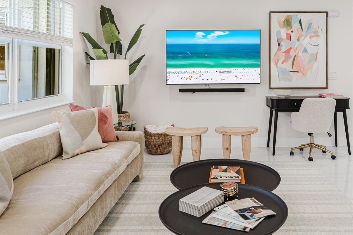 Light-filled living room with a 65" smart TV, sound bar, and subwoofer, featuring plush designer seating, neighborhood views, curated games, and reading materials—perfect for relaxing, entertaining, and family movie nights in West Palm Beach.