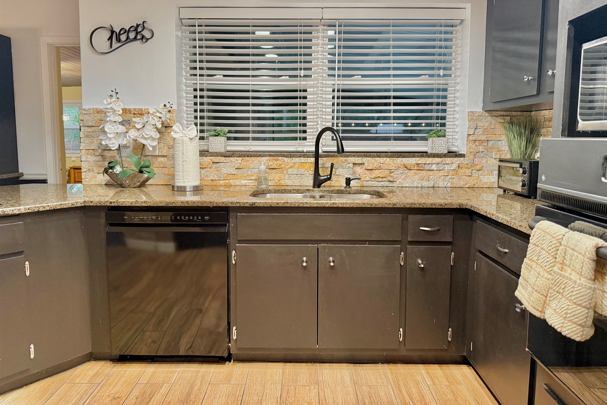 This sleek, dark-toned kitchen makes a stylish statement with black cabinetry, warm stone backsplash, and wood-look floors—perfect for whipping up a feast or morning coffee.