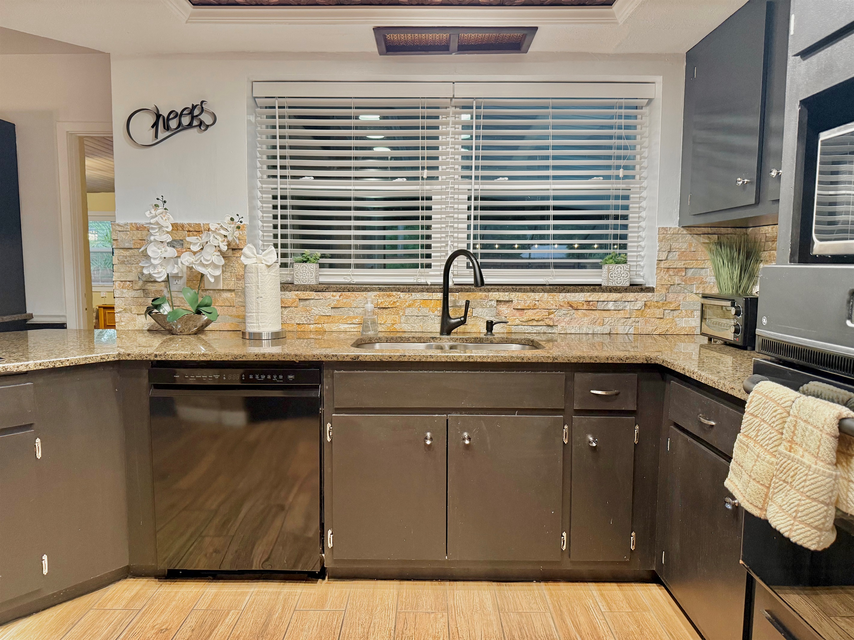 This sleek, dark-toned kitchen makes a stylish statement with black cabinetry, warm stone backsplash, and wood-look floors—perfect for whipping up a feast or morning coffee.