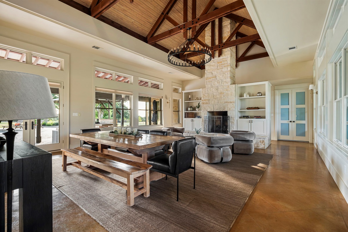 A spacious and inviting living area featuring high ceilings with exposed beams, a large dining table with benches, a cozy fireplace, and ample natural light.