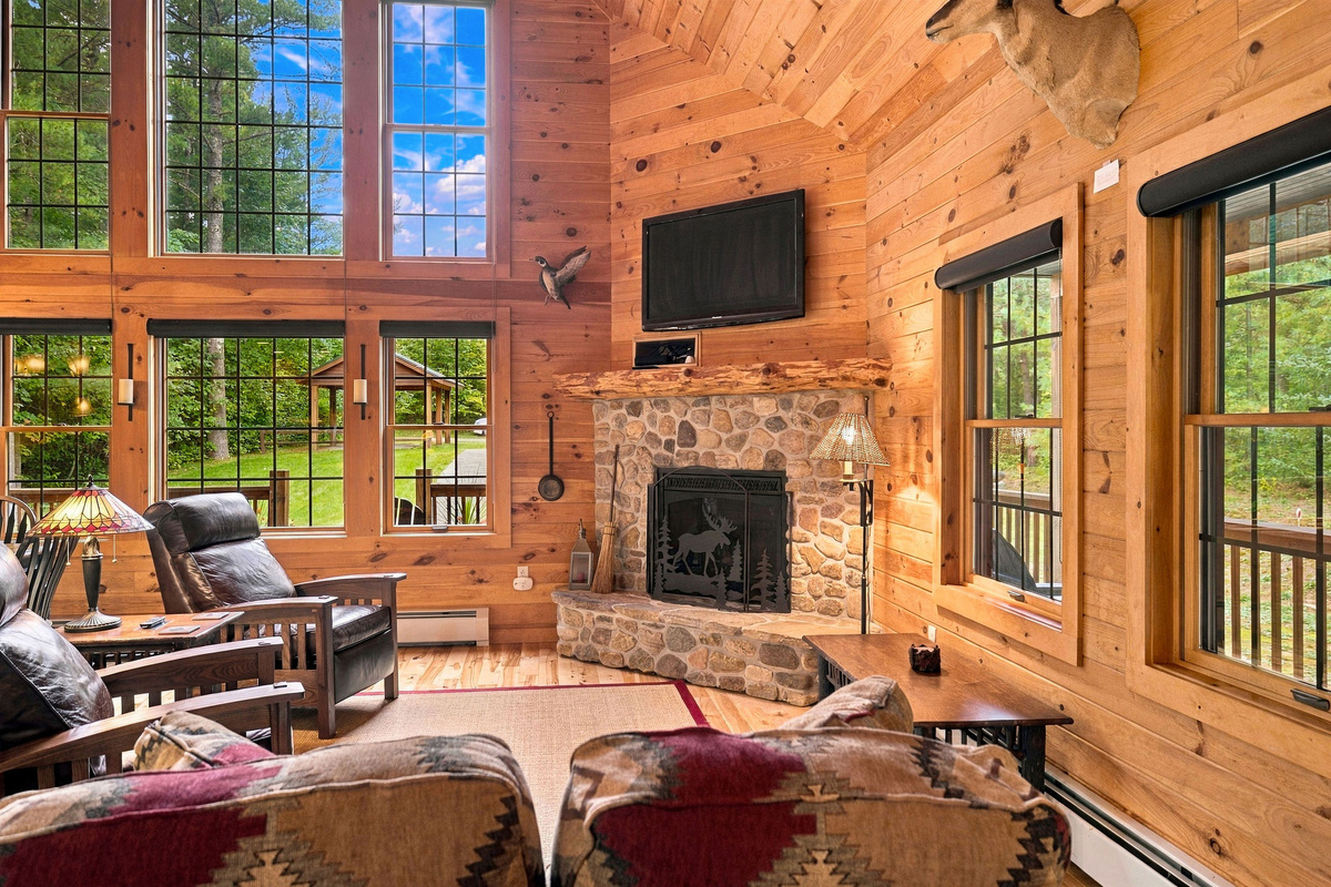 Soaring windows, warm wood interiors, large smart TV, and a stone fireplace create an inviting Adirondack great room.