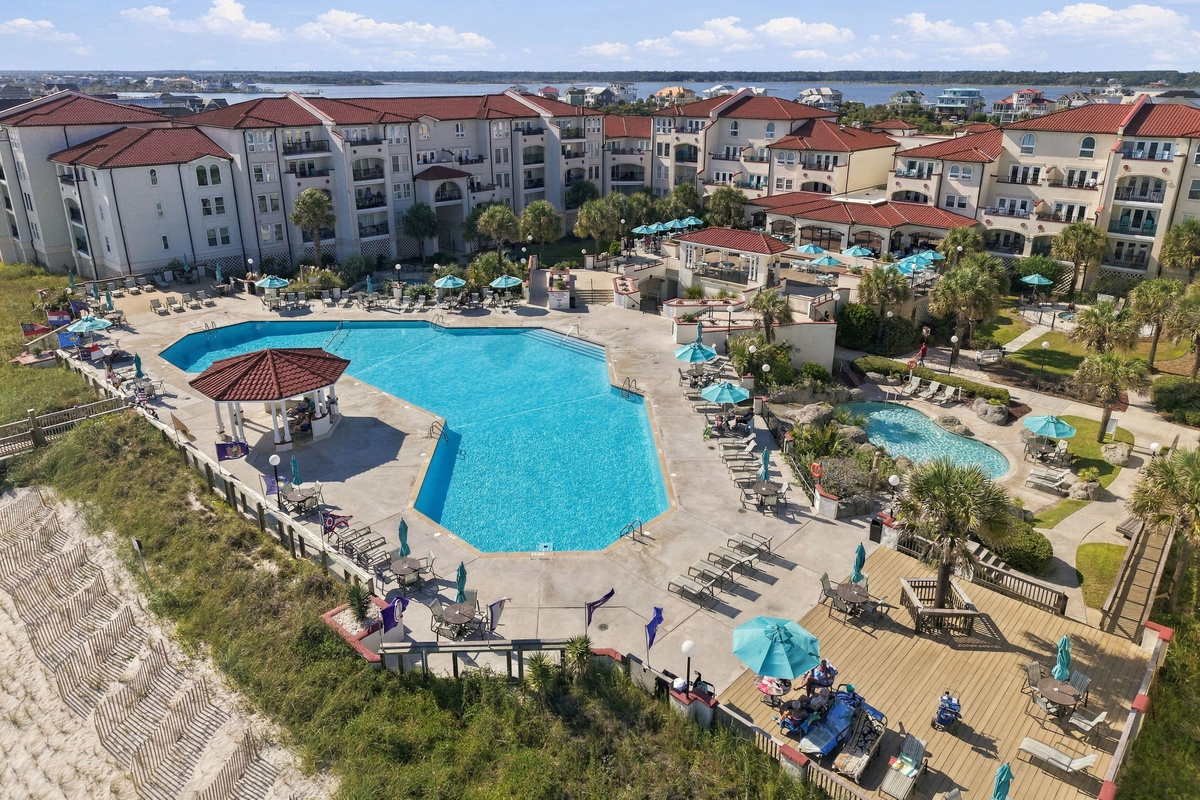 Outdoor pool deck featuring a swimming basin, shaded gazebo, wooden sun deck, lounge chairs, and palm-planted grounds alongside the coastal dunes.