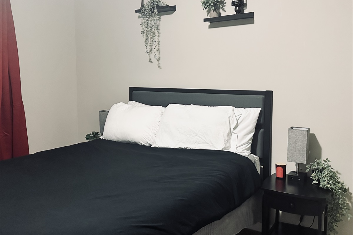 A cozy bedroom featuring a black bedspread draped over a neatly made bed with white pillows, adjacent to a black nightstand holding a small lamp and a candle, with decorative plants above and next to the bed, and a deep red curtain on one wall.