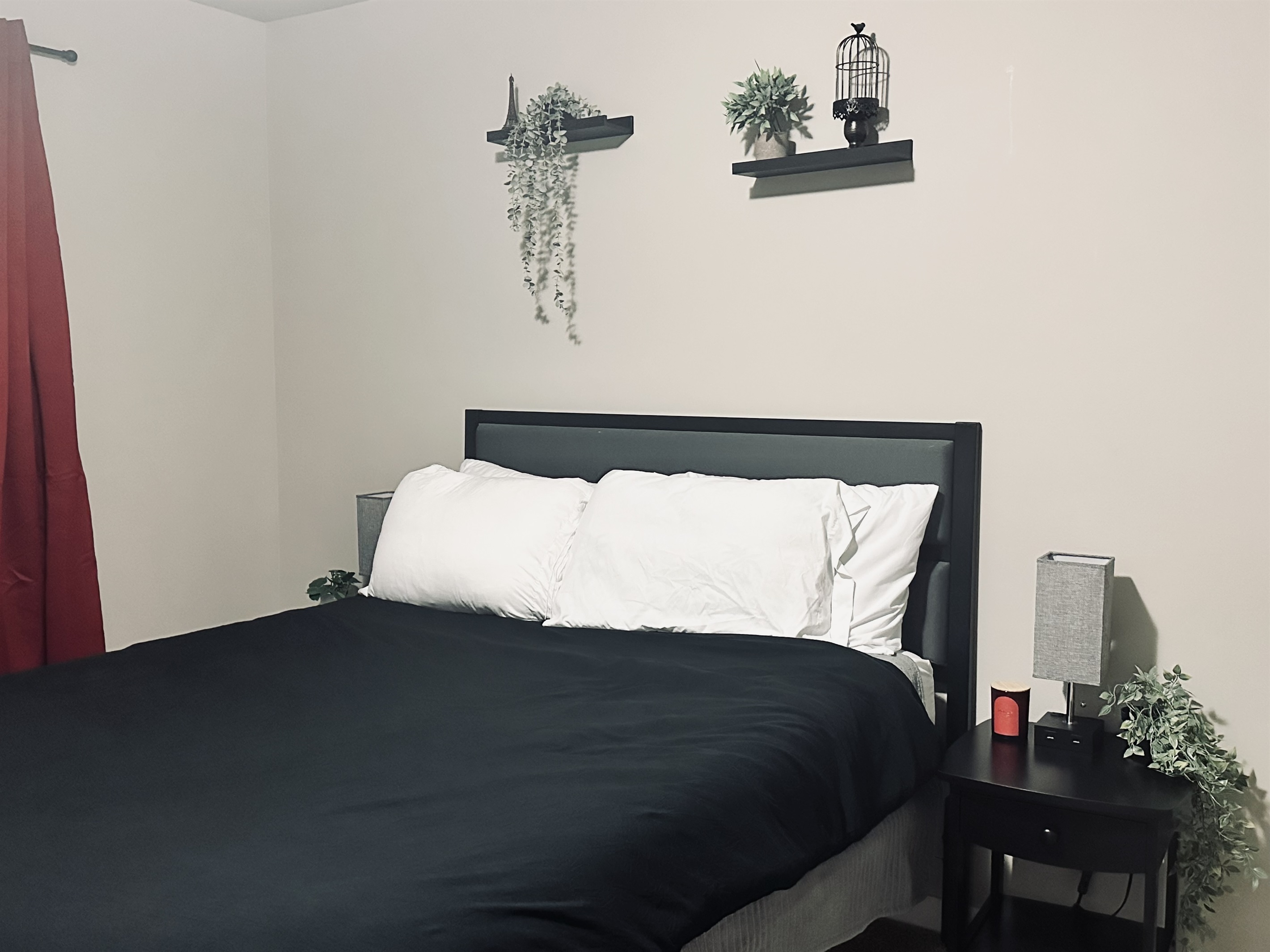 A cozy bedroom featuring a dark bedspread with white pillows, black bedside table with a lamp and a small decorative plant, red curtains, and wall shelves adorned with greenery and a decorative birdcage.