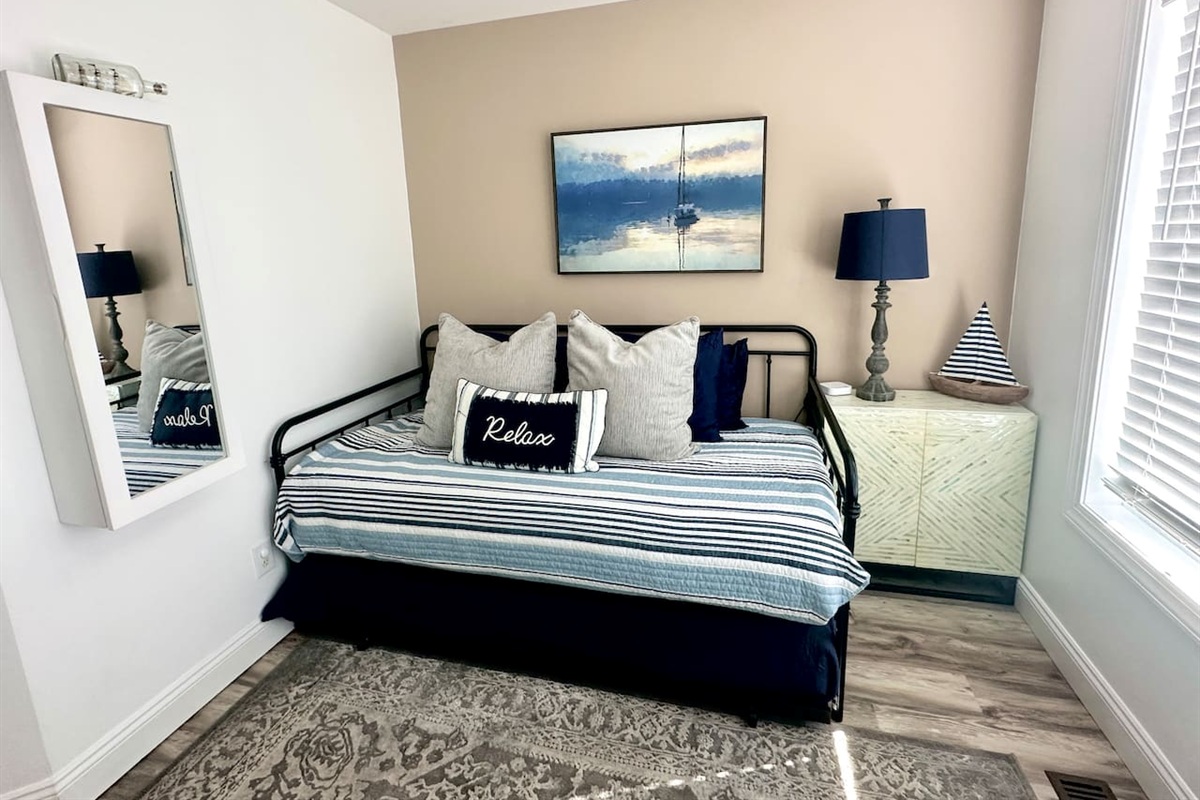 This versatile sunroom features a black metal daybed with nautical pillows and a striped coverlet. The space includes a large patterned rug, a light-green storage cabinet with a sailboat decor piece, and a wall-mounted mirror.