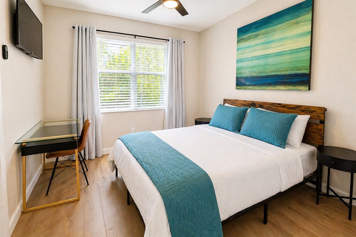 Bright and inviting bedroom with a comfortable bed, fresh linens, and modern coastal décor. Includes a dedicated workspace, wall-mounted TV, and plenty of natural light—perfect for both relaxing and getting things done.