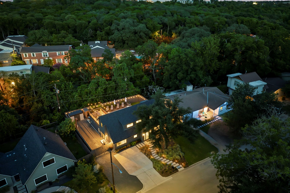 An aerial nighttime view of a modern home with a well-lit driveway and a spacious deck for entertaining, nestled within a lush, tree-filled neighborhood.