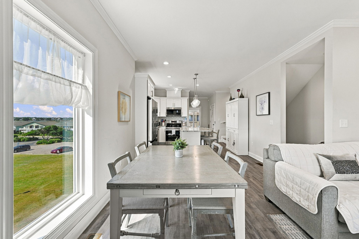 Sunlit dining area featuring a whitewashed wooden dining table, neutral chair seating, and large picture windows framing scenic neighborhood views, with an effortless connection to the modern kitchen beyond.   