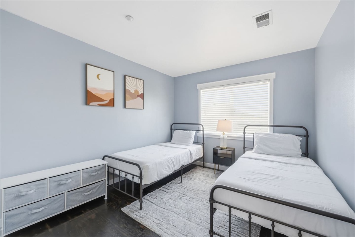Bedroom 2 with twin beds, fresh bed linens, room-darkening shades, and clothing storage. Comfortable setup for kids or traveling companions.