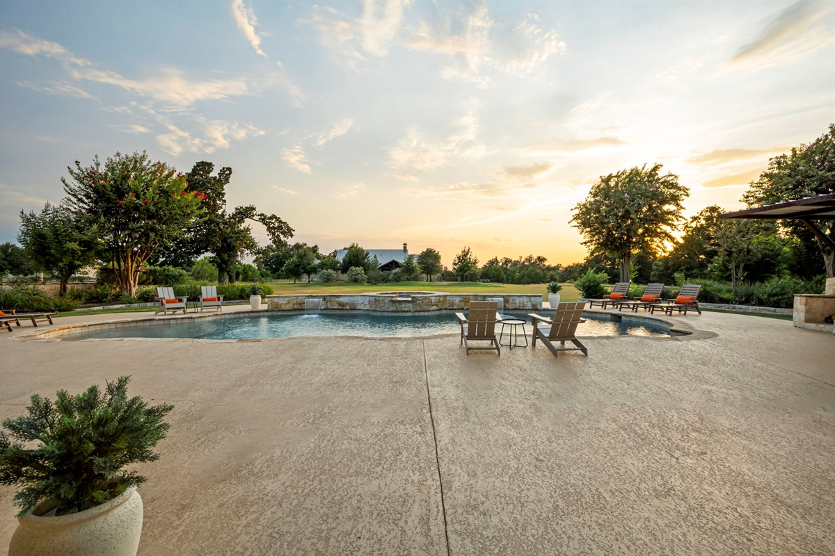 A luxurious Hill Country home featuring a sprawling outdoor living area with a resort-style pool, hot tub, and ample lounging space, set against a clear blue sky.