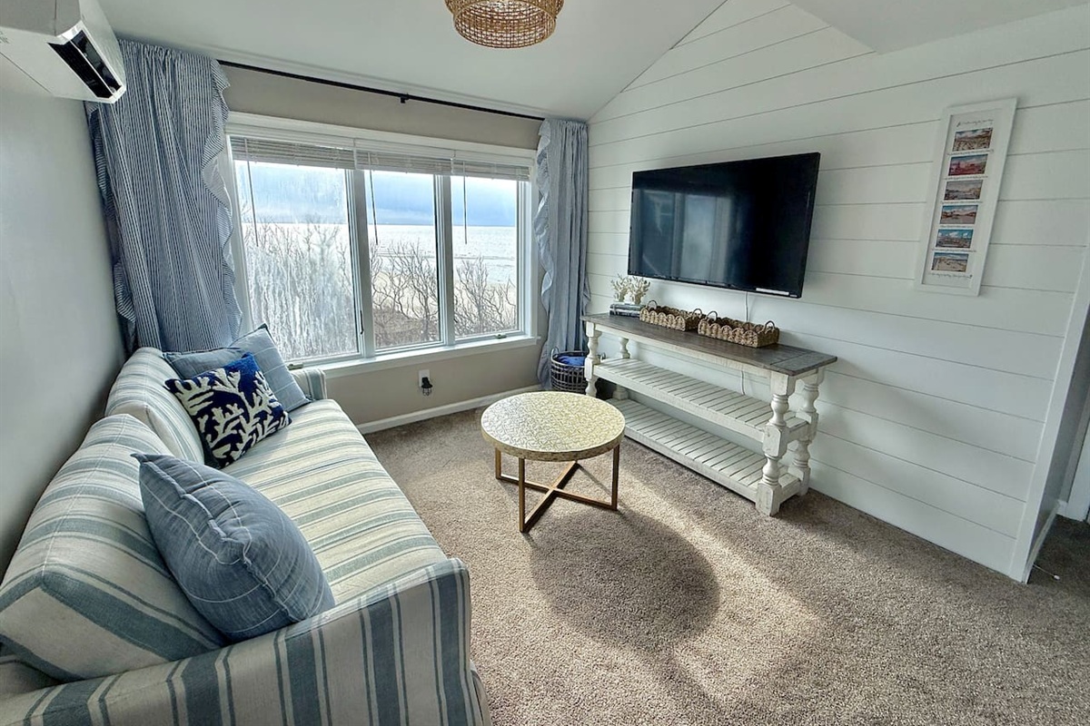 Unwind in this cozy, coastal-inspired sitting area that features comfortable striped seating, a rustic console table with a wall-mounted television, and large windows offering relaxing views for the perfect vacation escape.