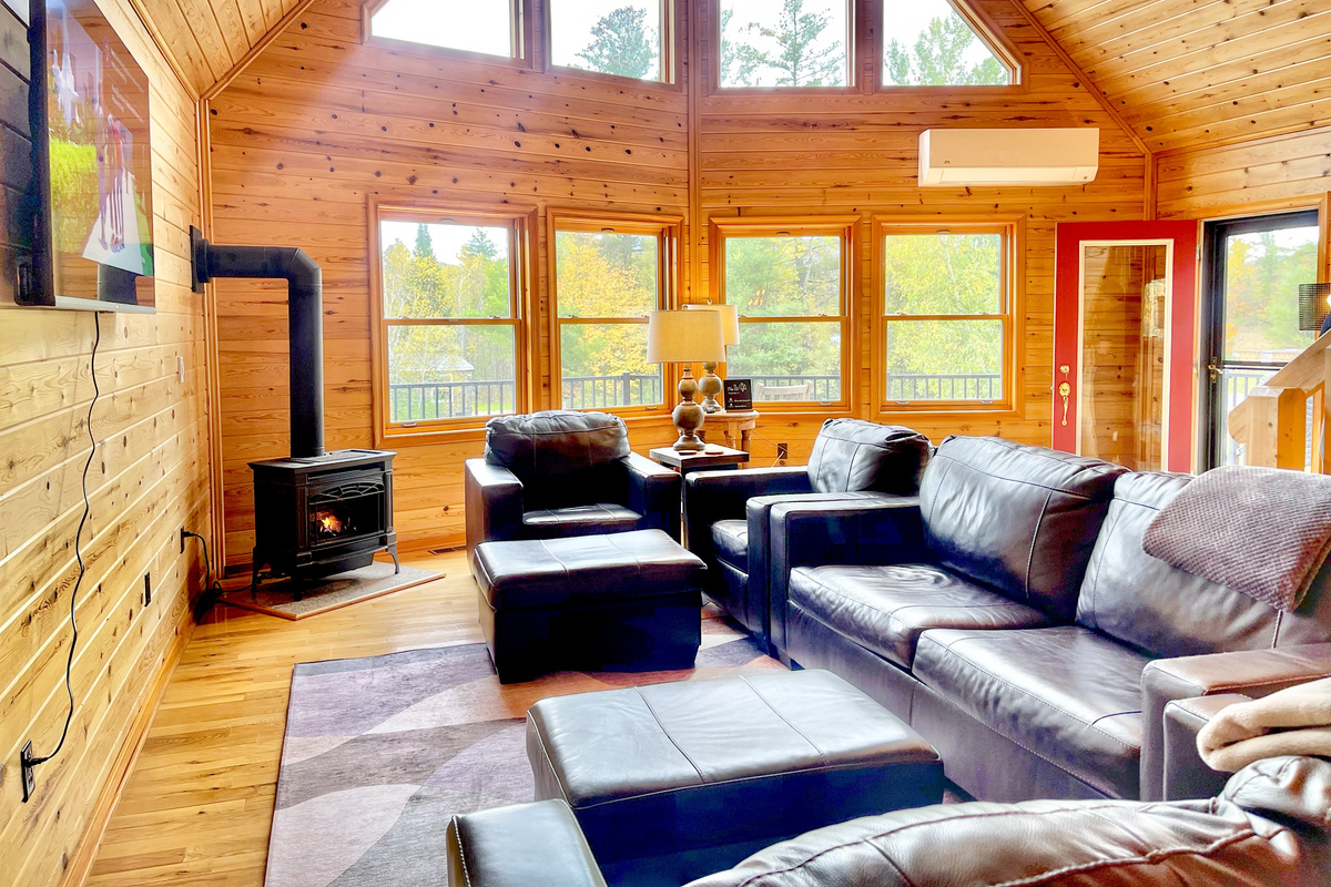 Unwind in the spacious living room, featuring comfortable leather seating, a cozy gas stove, a 60-inch Smart TV, and expansive windows overlooking the surrounding trees.