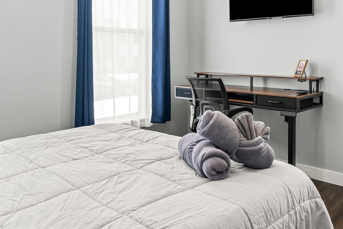 Bedroom workspace angle featuring a desk, ergonomic chair, wall-mounted TV, and natural light.