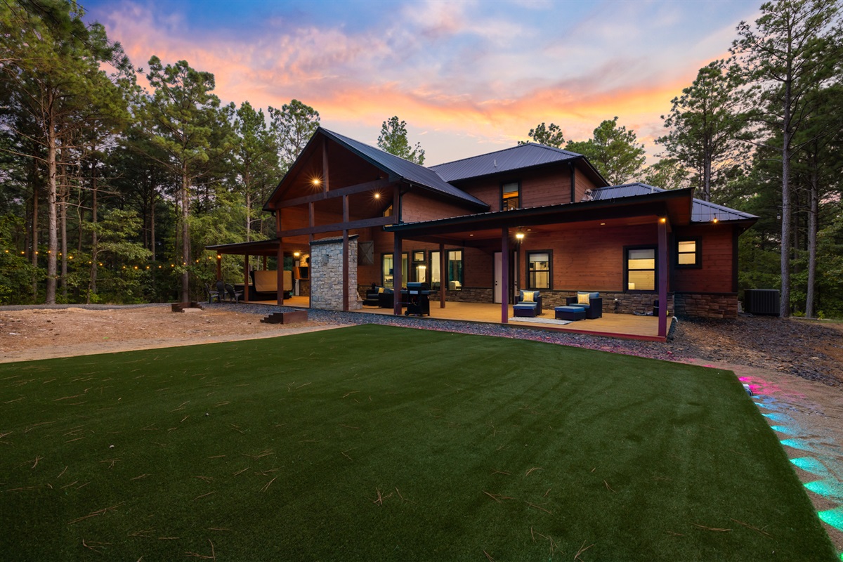 Exterior view of Red River Lodge featuring a modern cabin surrounded by tall pines in Hochatown. The home showcases a spacious outdoor area with LED-lit turf, hot tub, firepit, and patio seating, offering a perfect blend of luxury, comfort, and nature.
