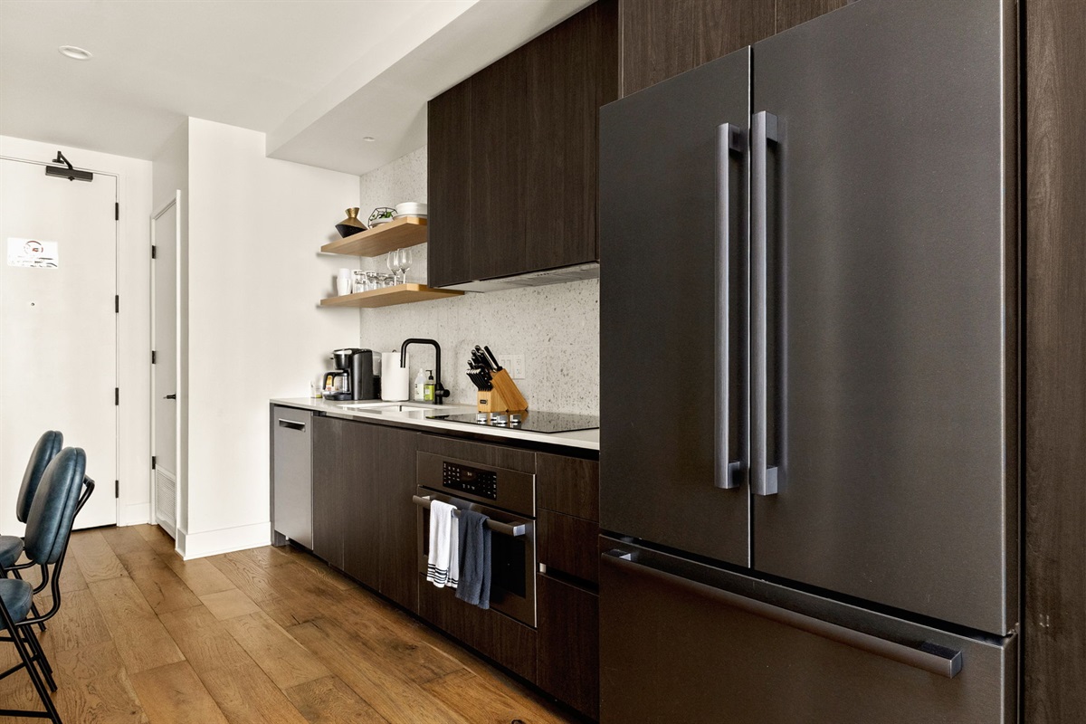 A modern kitchen featuring dark wood cabinetry, stainless steel appliances, and a light-colored speckled countertop with a matching backsplash. The space is equipped with a full-sized refrigerator, dishwasher, and stovetop, and is accented with open shelvi