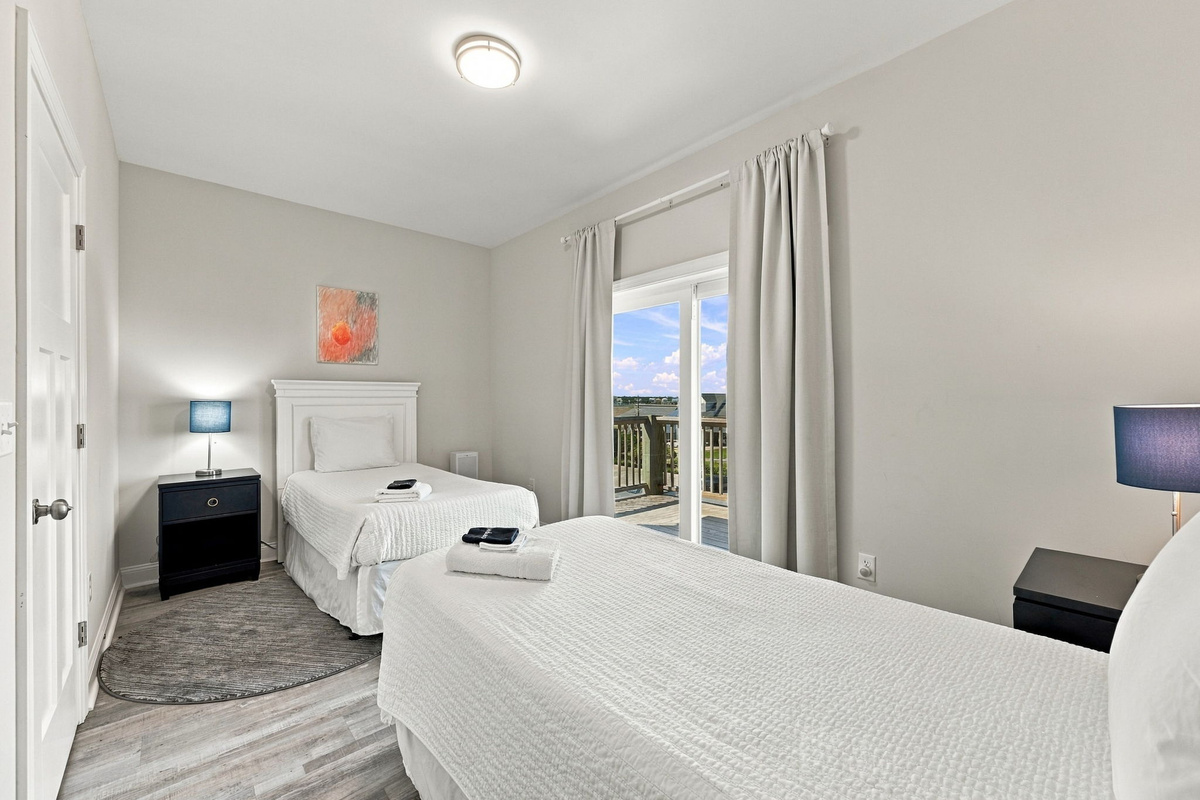 Light-filled guest bedroom featuring twin beds dressed in clean white linens, dark nightstands with stylish accent lamps, soft neutral walls, and a sliding glass door offering direct access to an outdoor deck area.   