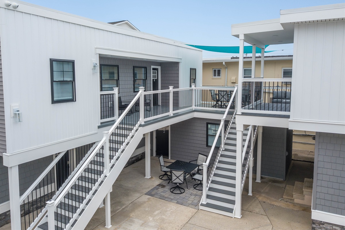 Modern beach-style condo with spacious shared patio, outdoor seating, and shaded upper decks - perfect for relaxing after a day by the shore. Clean, coastal design and easy access via dual staircases.