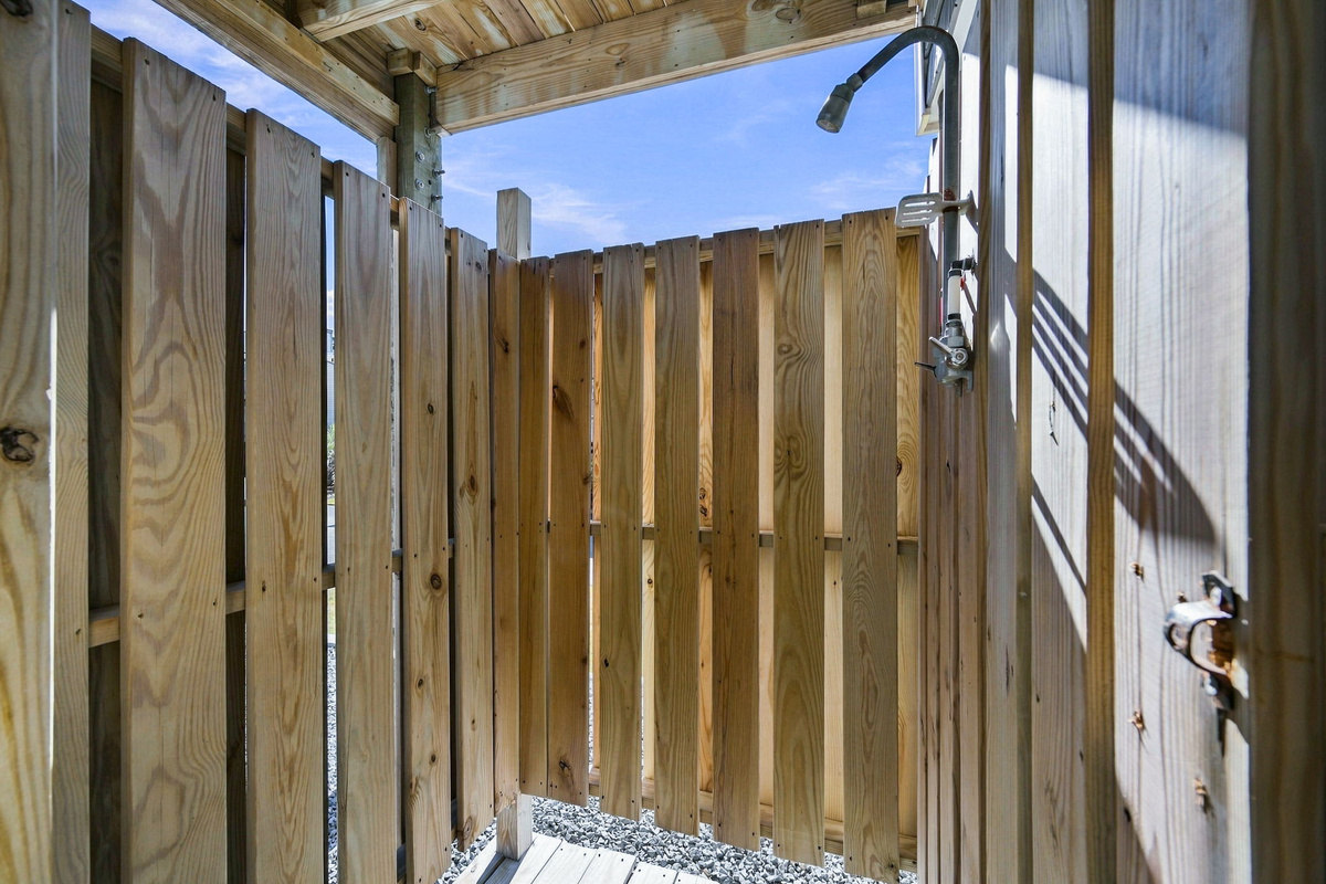 Enclosed ground-level outdoor shower featuring vertical wooden privacy slats, a shower head fixture, and a latched door for convenient post-beach cleanup.   