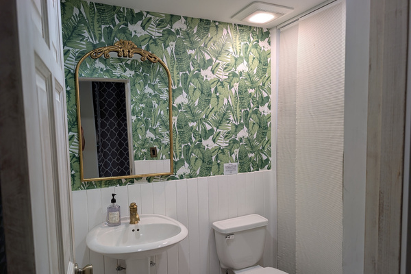 Social half-bath at Villa Coral featuring vibrant green tropical leaf wallpaper, white vertical shiplap wainscoting, a pedestal sink, and a vintage gold arched mirror in Dorado, Puerto Rico.
