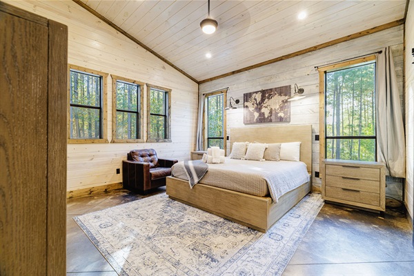 Cozy rustic bedroom with a modern farmhouse design, featuring a plush king-size bed, wood-paneled walls, large windows with forest views, and warm lighting. Perfect for a cabin retreat or vacation rental.