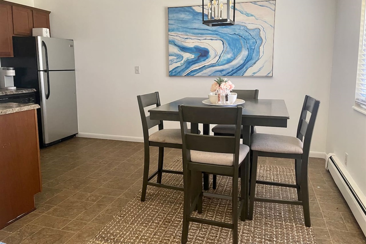 Bright, modern dining area with seating for four and eye-catching coastal artwork. Enjoy meals, game nights, or remote work under stylish lighting with natural light streaming in—an inviting space for connection and comfort during your stay.