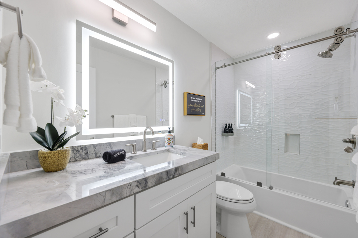 A bright full bathroom includes an illuminated mirror, modern vanity, and glass-enclosed tub/shower for easy daily routines.