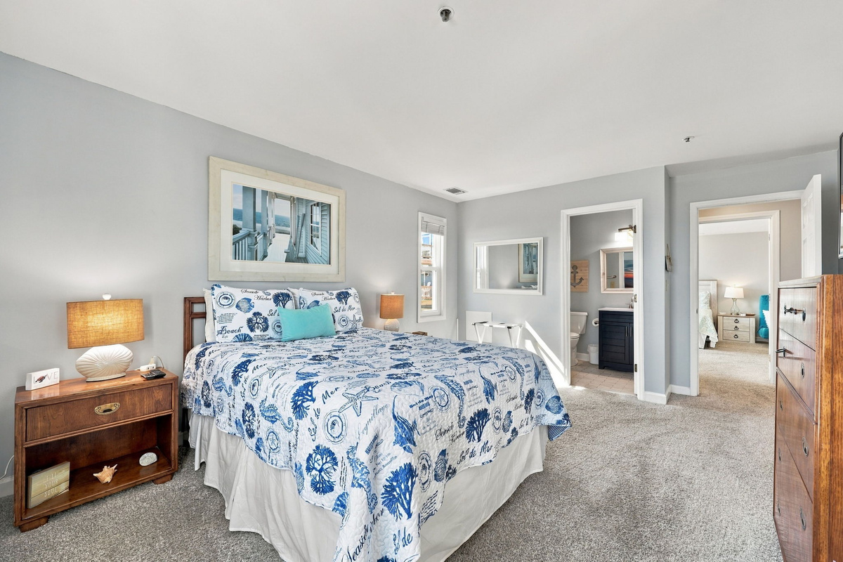 Well-appointed guest bedroom featuring nautically patterned bedding, warm wood nightstands and dressers, framed coastal art, and direct access to an en-suite bathroom.   