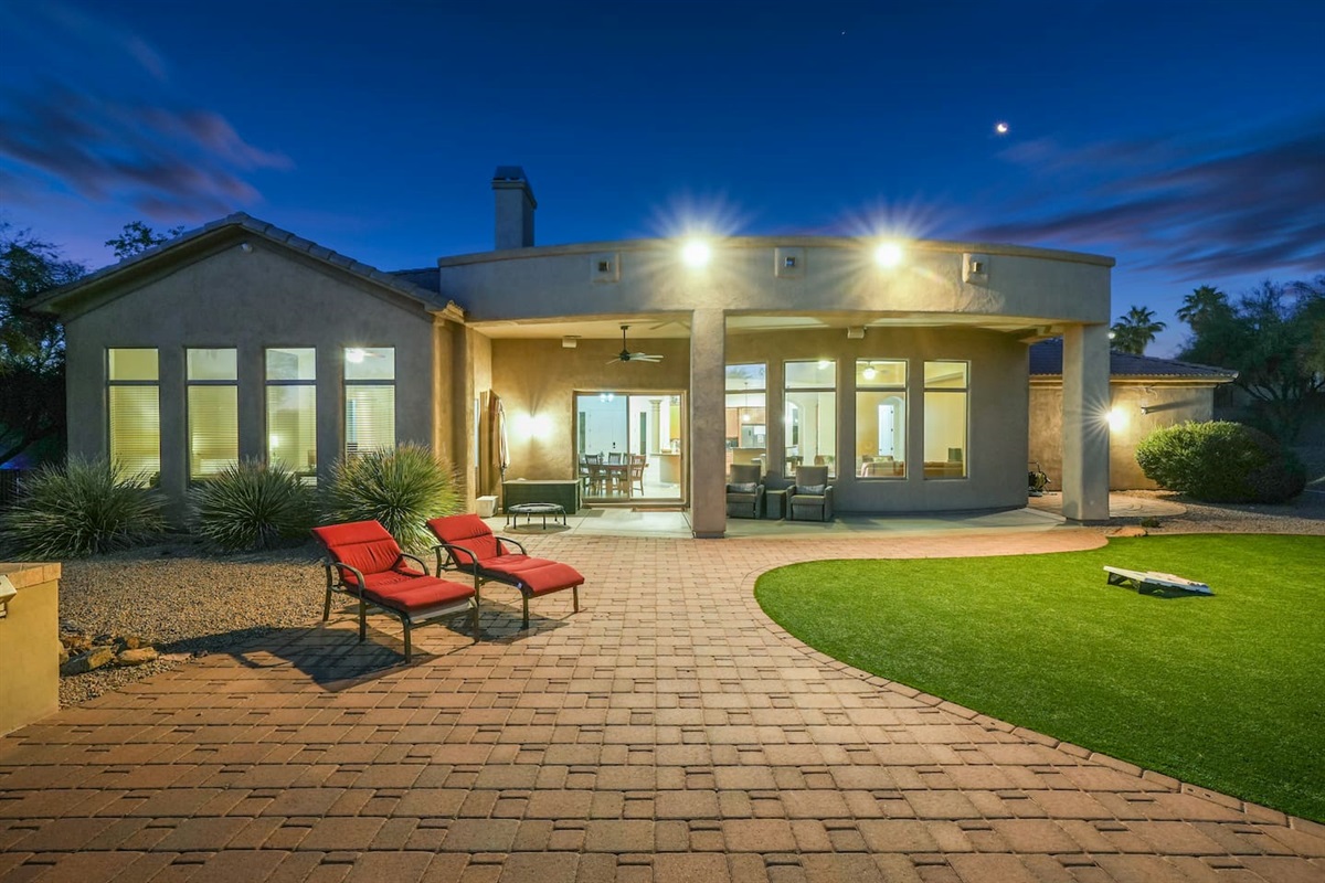 Evening backyard ambiance with soft lighting, red loungers, and a warm glow flowing from the home for nighttime relaxation.