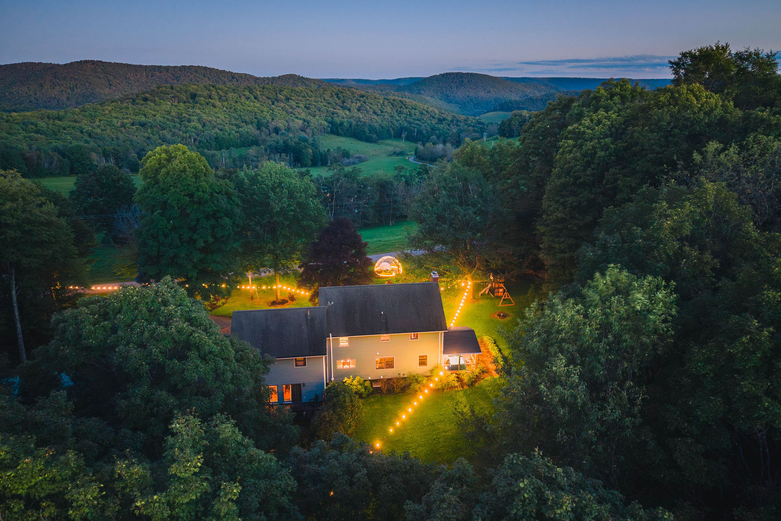 💡 The house lit up at dusk, dome and swing set on the lawn.