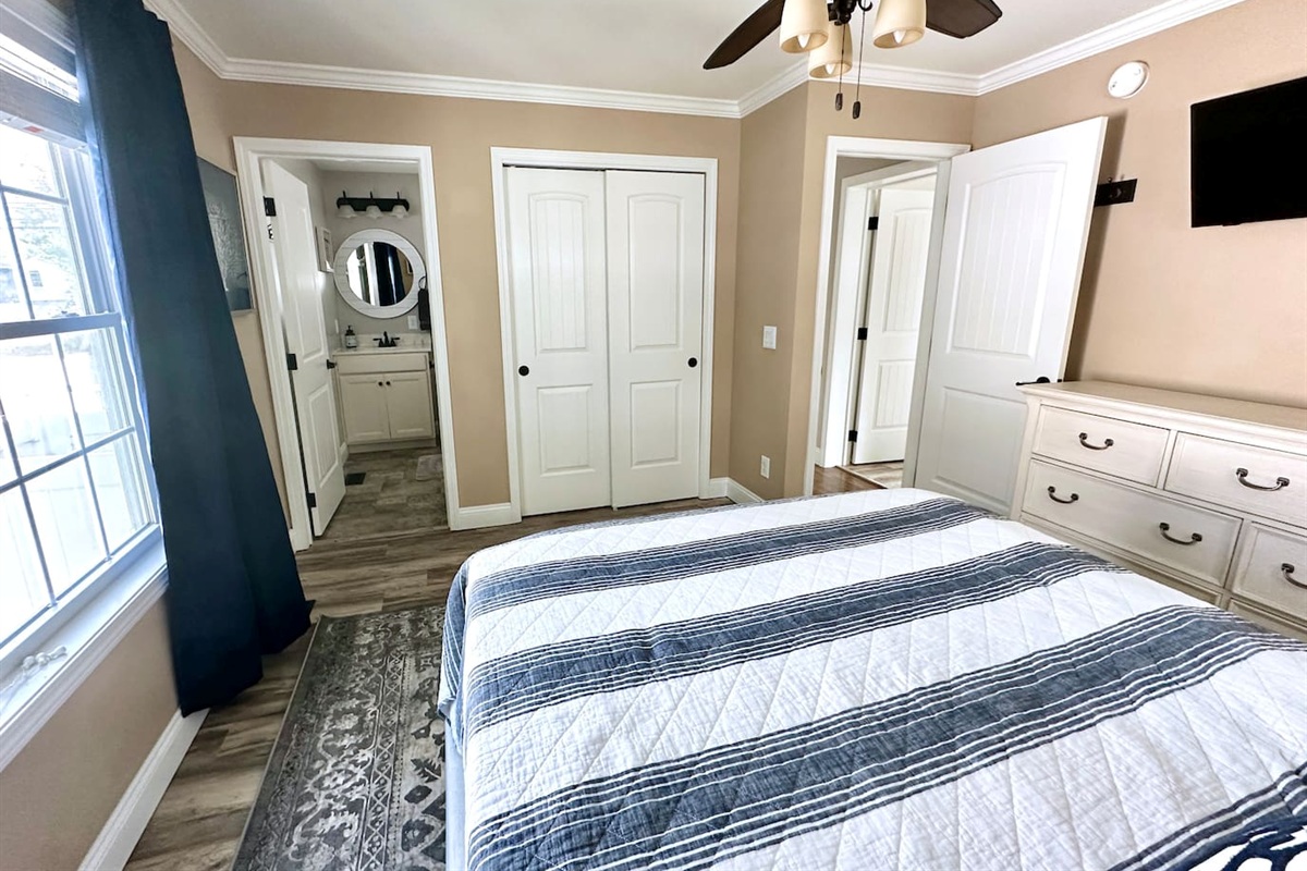 This bedroom provides the convenience of an en-suite bathroom and a large closet. The space is thoughtfully arranged with a dresser, wall-mounted TV, and hardwood-style flooring.