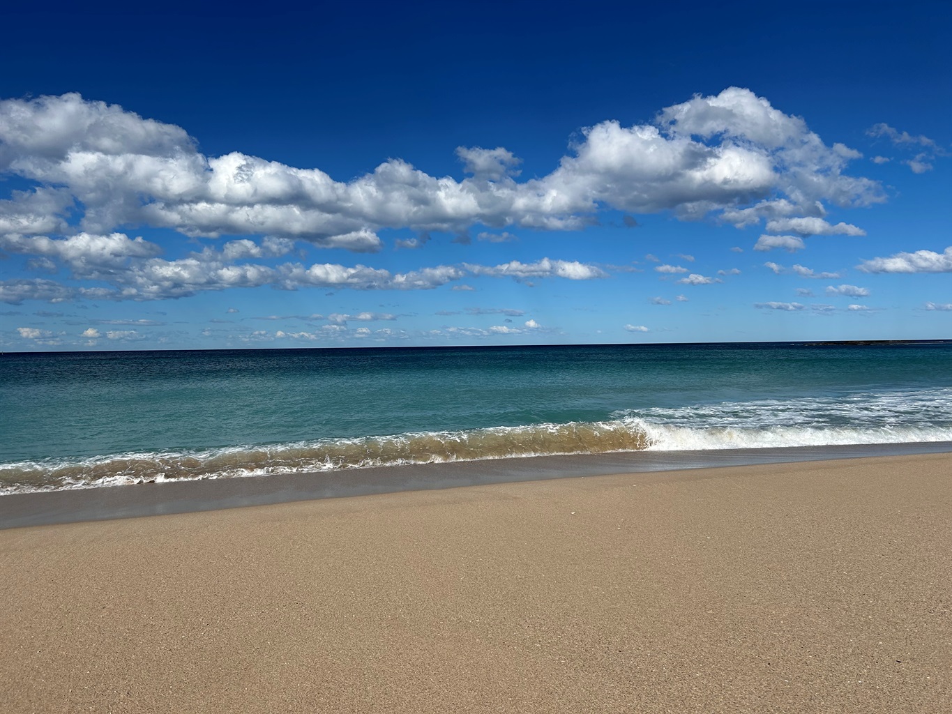 Blue sky with wet sand in the foreground and unusually small waves and shades of green and blue ocean in the background