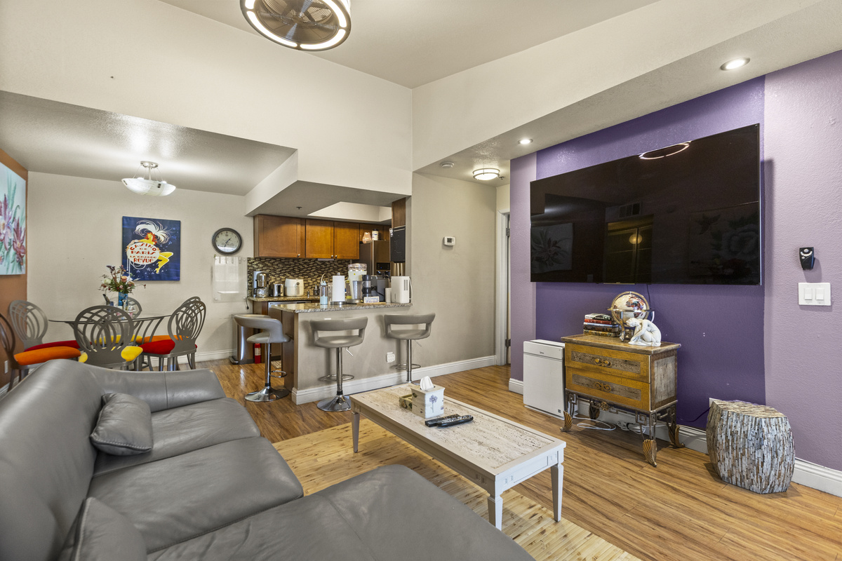 Living room with gray sofa seating, wall-mounted Smart TV, bar seating, and the kitchen and dining area close by.
