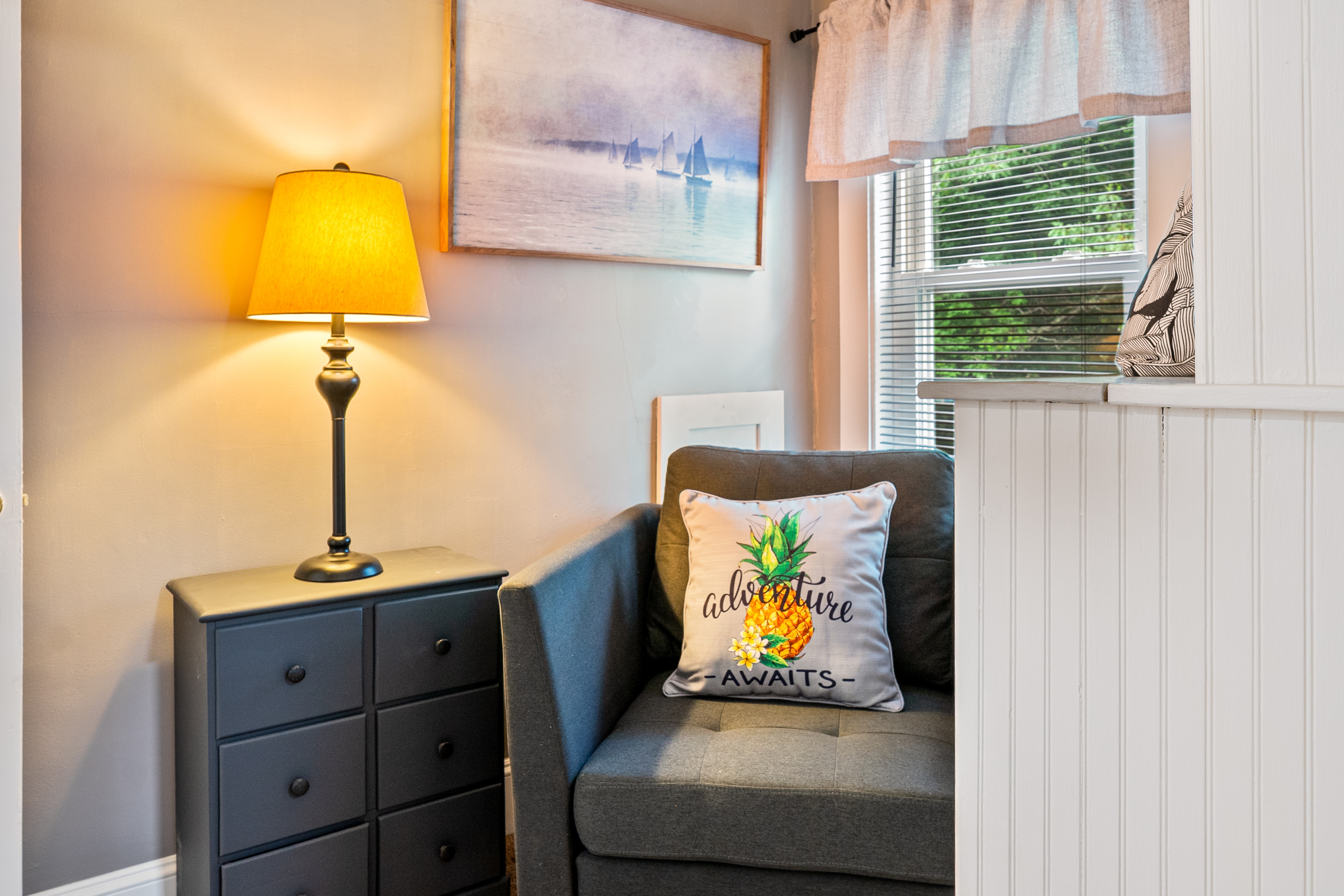Peeking out from the queen bedroom, this perspective highlights the inviting warmth of The Pebble Nook. The cozy armchair setup is complete with a lit reading lamp, functional clothing storage drawers, a leafy window view, and misty sailboat artwork.