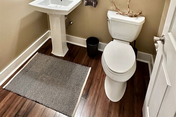 This classic powder room features a white pedestal sink and dark wood-style flooring. The neutral walls and simple patterned rug create a clean, inviting space for guests.