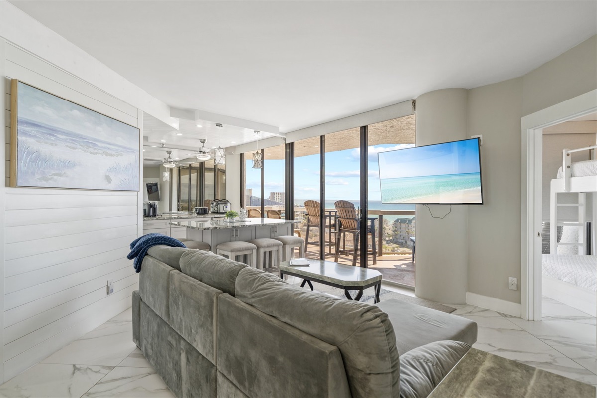 This is the view from the sofa. The Gulf fills the glass from corner to corner, the balcony is right there, and the kitchen is just behind you. This condo was built around this exact moment.