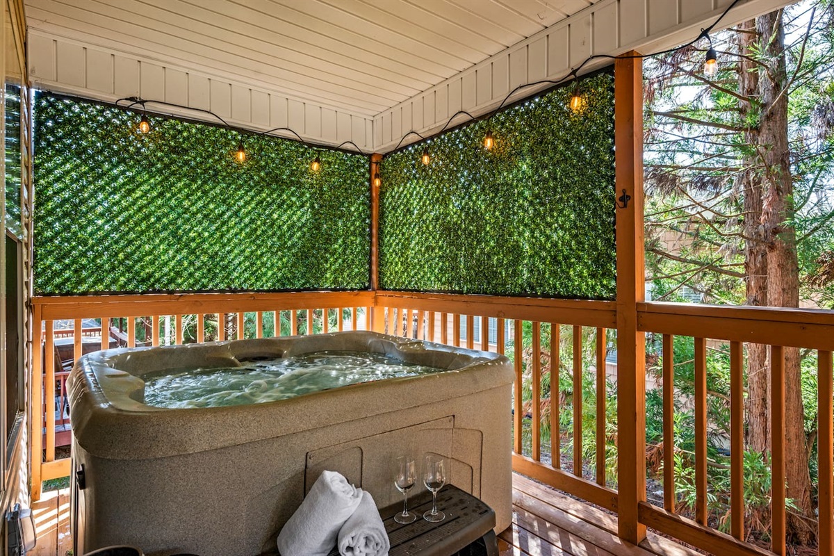 This inviting covered deck features a private hot tub surrounded by lush greenery panels for added privacy. String lights create a cozy, ambient glow—perfect for unwinding in the evening. With soft towels, wine glasses, and peaceful wooded views, this spac