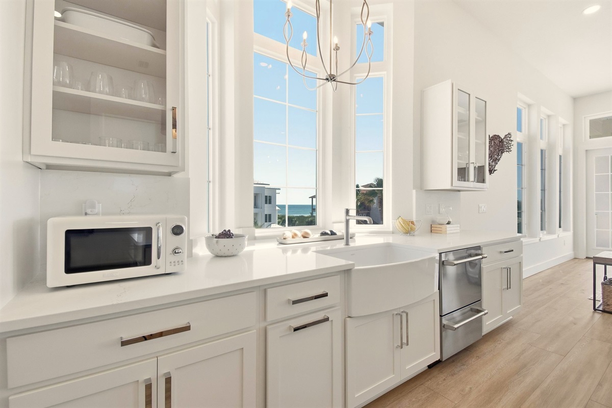 Morning light pours through soaring windows as you rinse sandy shells in the farmhouse sink, brew coffee, and watch a ribbon of ocean shimmer beyond the rooftops. The whole kitchen feels like a calm exhale before the day begins.
