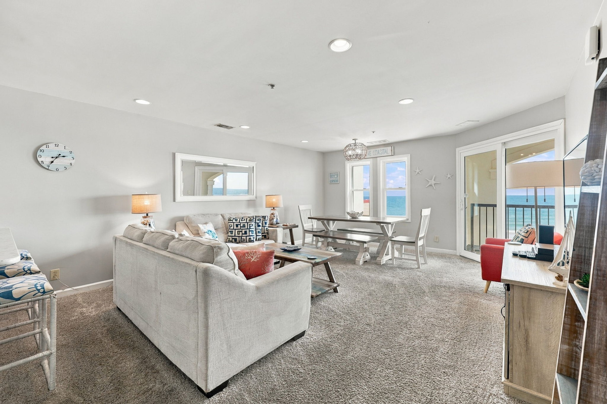 Bright and airy main living sanctuary featuring plush neutral carpeting, a comfortable neutral sofa, dedicated dining area, and wall-to-wall sliding glass doors framing captivating ocean views and leading out to a private balcony deck.   