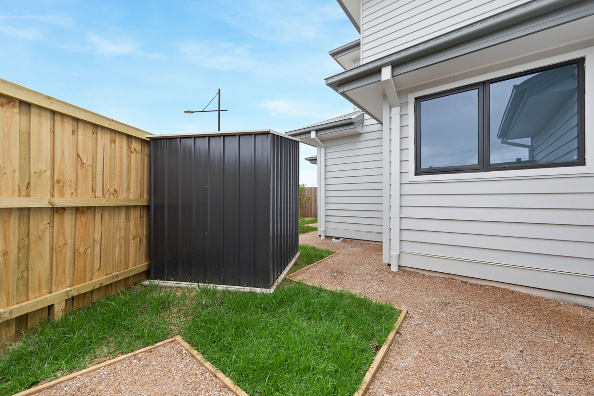 Side yard with a lawn strip, gravel path and a dark storage shed, enclosed by timber fencing.